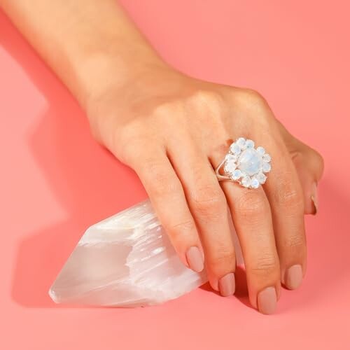 Hand with a silver ring featuring a blue gemstone, holding a clear crystal on a pink background.
